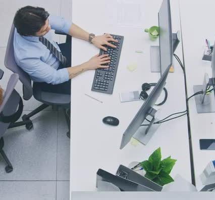 Aerial view of a bright business office showing a row of desks with diverse young professionals actively working on desktop computers and talking with each other.