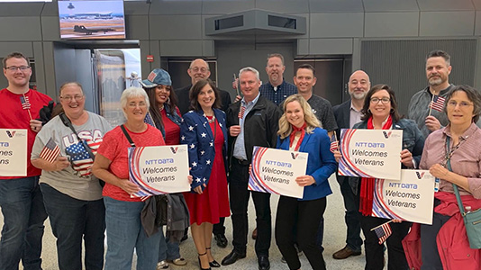 Group of people showcasing a billboard that reads "NTT DATA Welcomes Veterans"