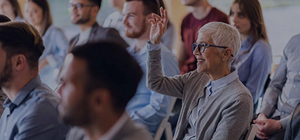 Happy senior businesswoman attending a seminar with her colleagues while raising her hand to ask a question